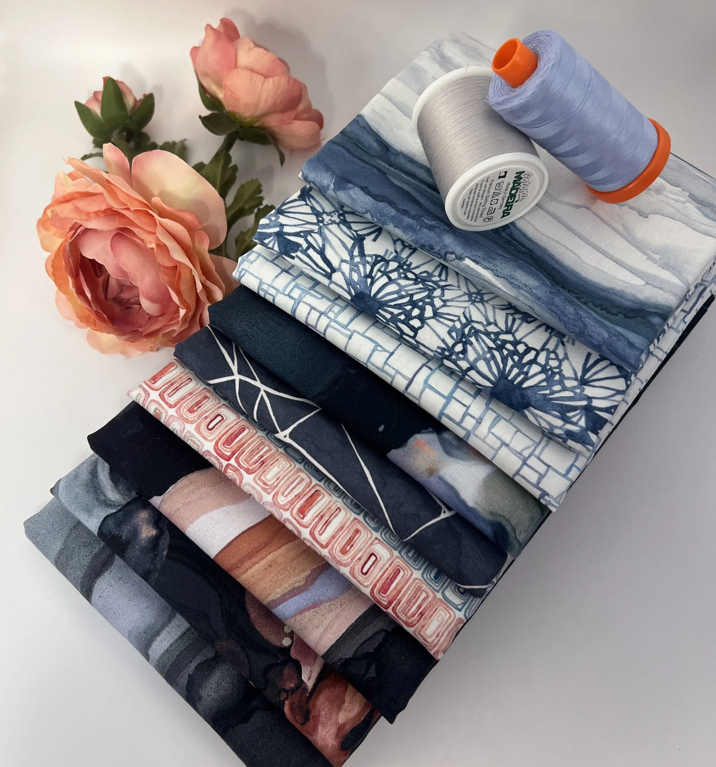 Close-up view of several folded fabric pieces with various blue, gray, black, and pattern designs, accompanied by three spools of thread in silver and light blue, placed on a white surface next to pink roses.