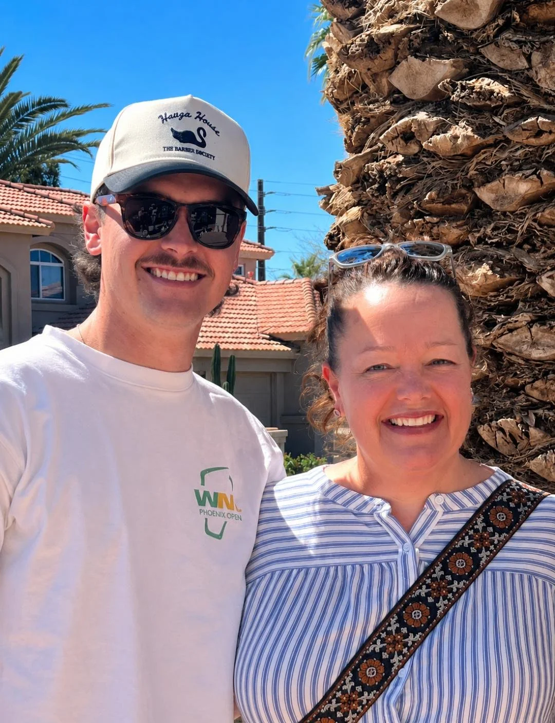 Kara and Kobe, founders of the company, smiling together outdoors, standing next to a large palm tree with a residential neighborhood in the background under a clear blue sky.