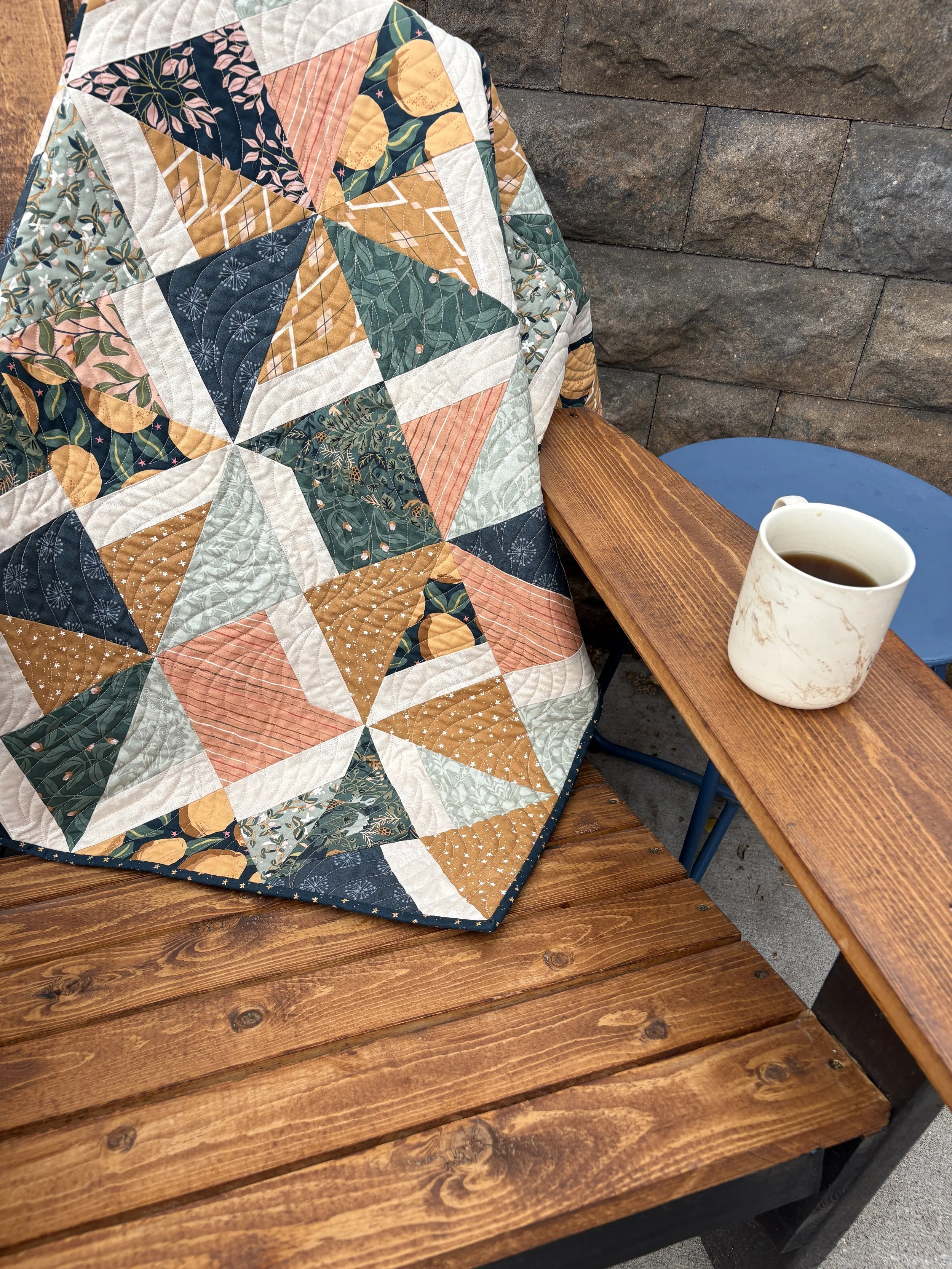 Close-up view of a quilt with various colorful patterns draped over a wooden bench. A white mug with coffee sits on the bench.