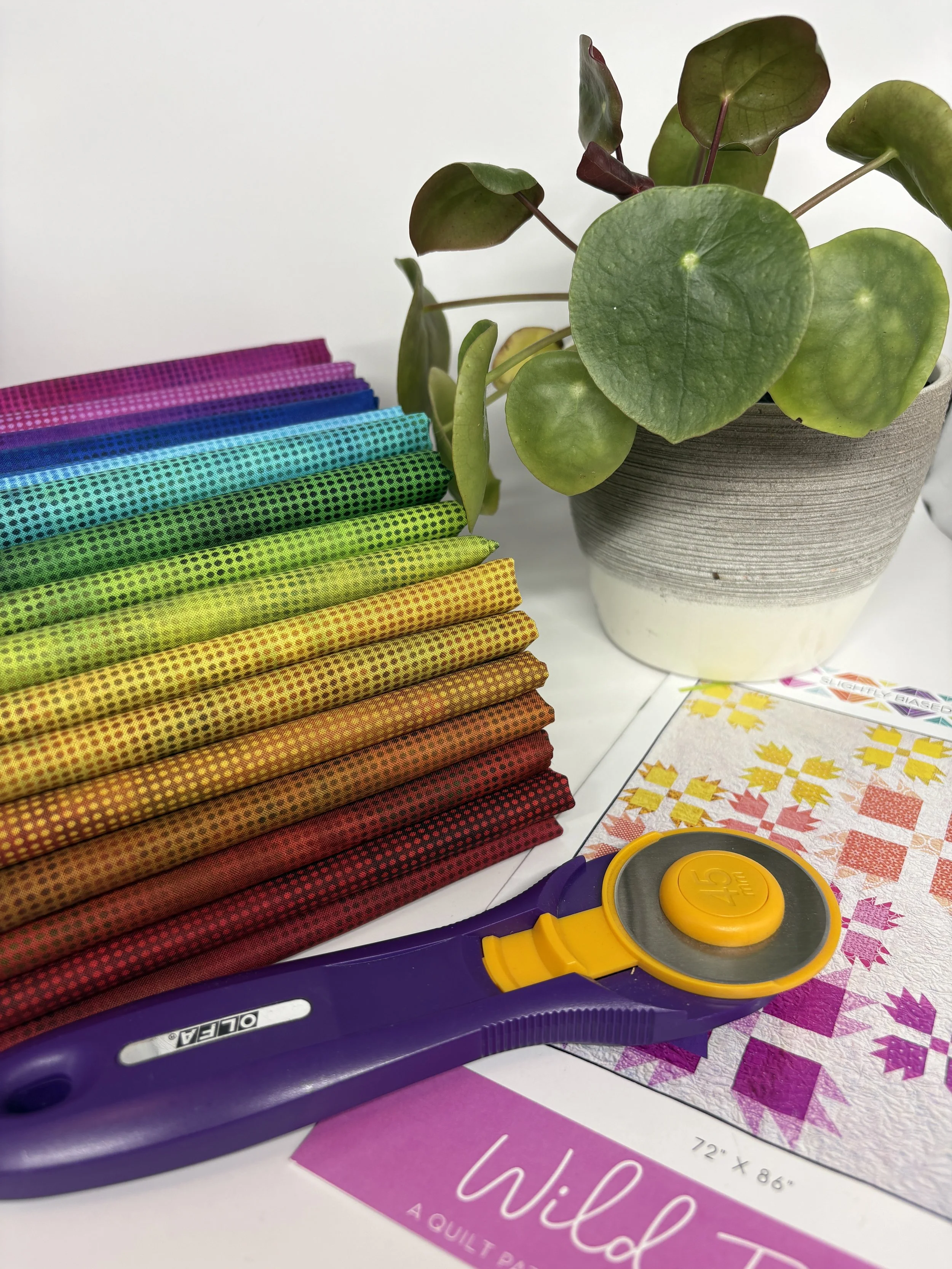 A stack of colorful fabric squares, a potted plant with large green leaves, a yellow rotary cutter, and a quilting pattern sheet.