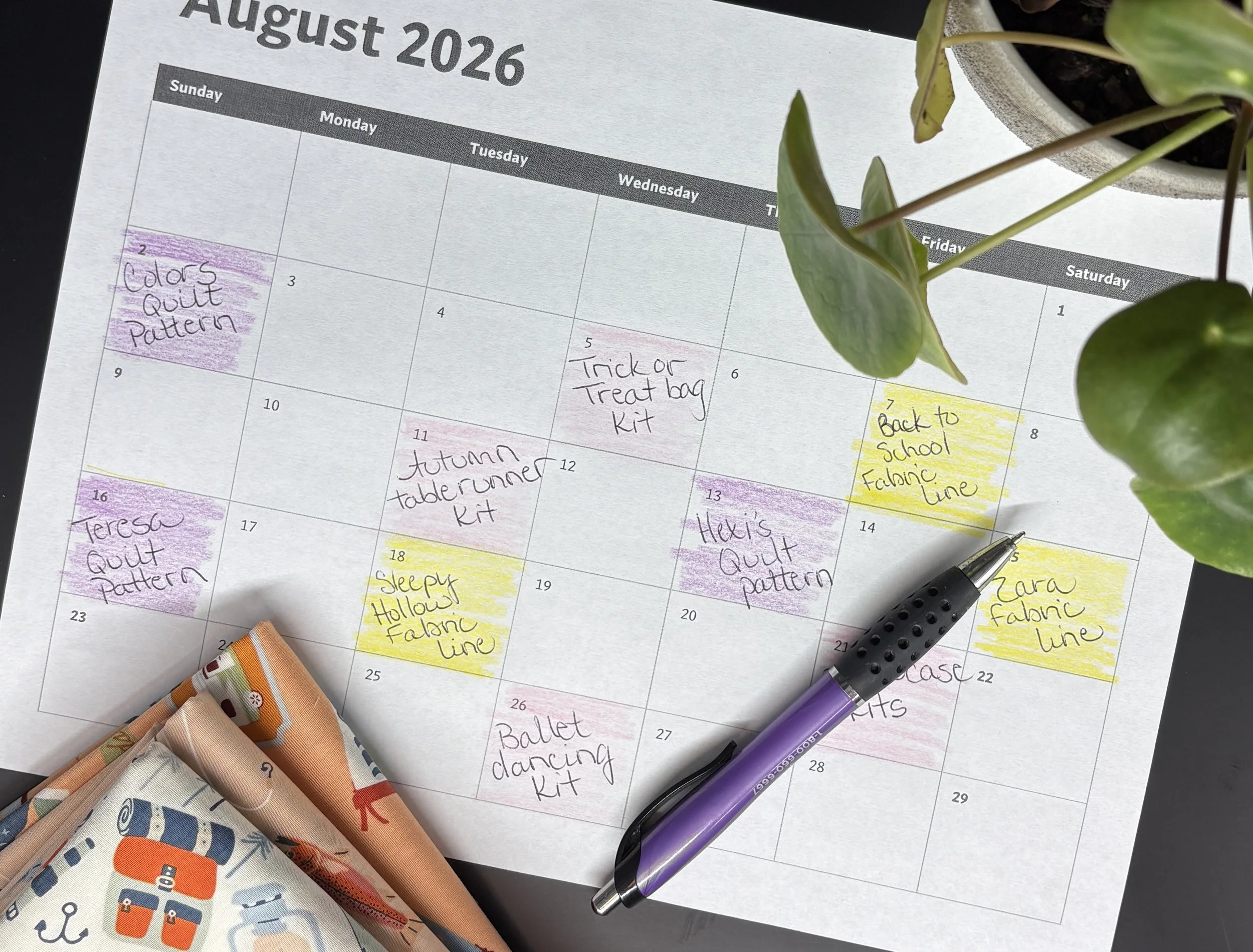 A calendar with handwritten notes in various colors. Visible notes include 'Colors Quilt Pattern,' 'Trick or Treat bag Kit,' 'Back to School Fabric Line,' 'Autumn Table Runner Kit,' 'Sleepy Hollow Fabric Line,' and 'Ballet dancing kit.' There are fabric samples, pens, and a potted plant partially visible around the calendar.