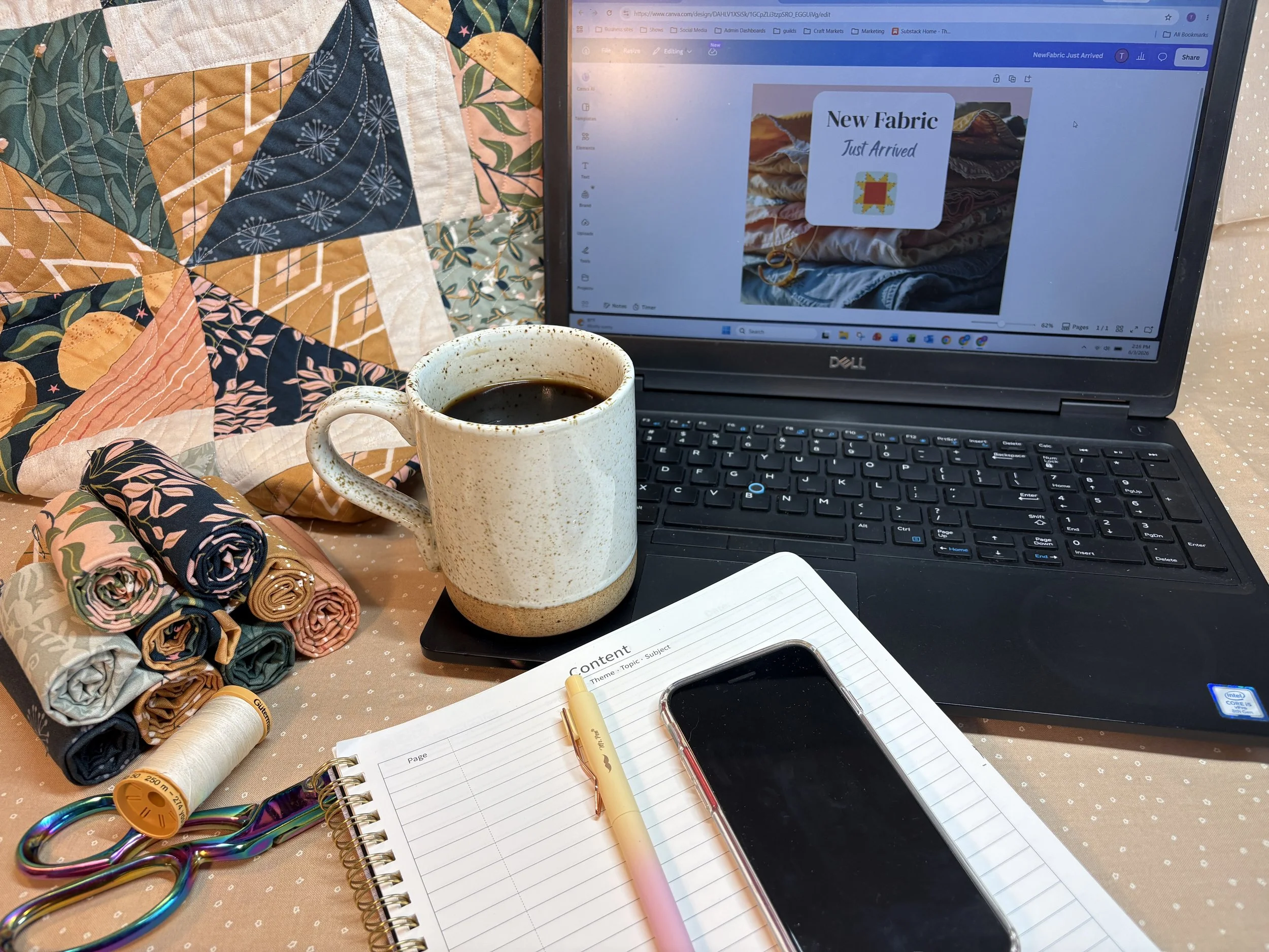 A workspace with a fabric quilt, a cup of coffee, sewing supplies, a notebook, a pen, a smartphone, and a laptop displaying a design project.