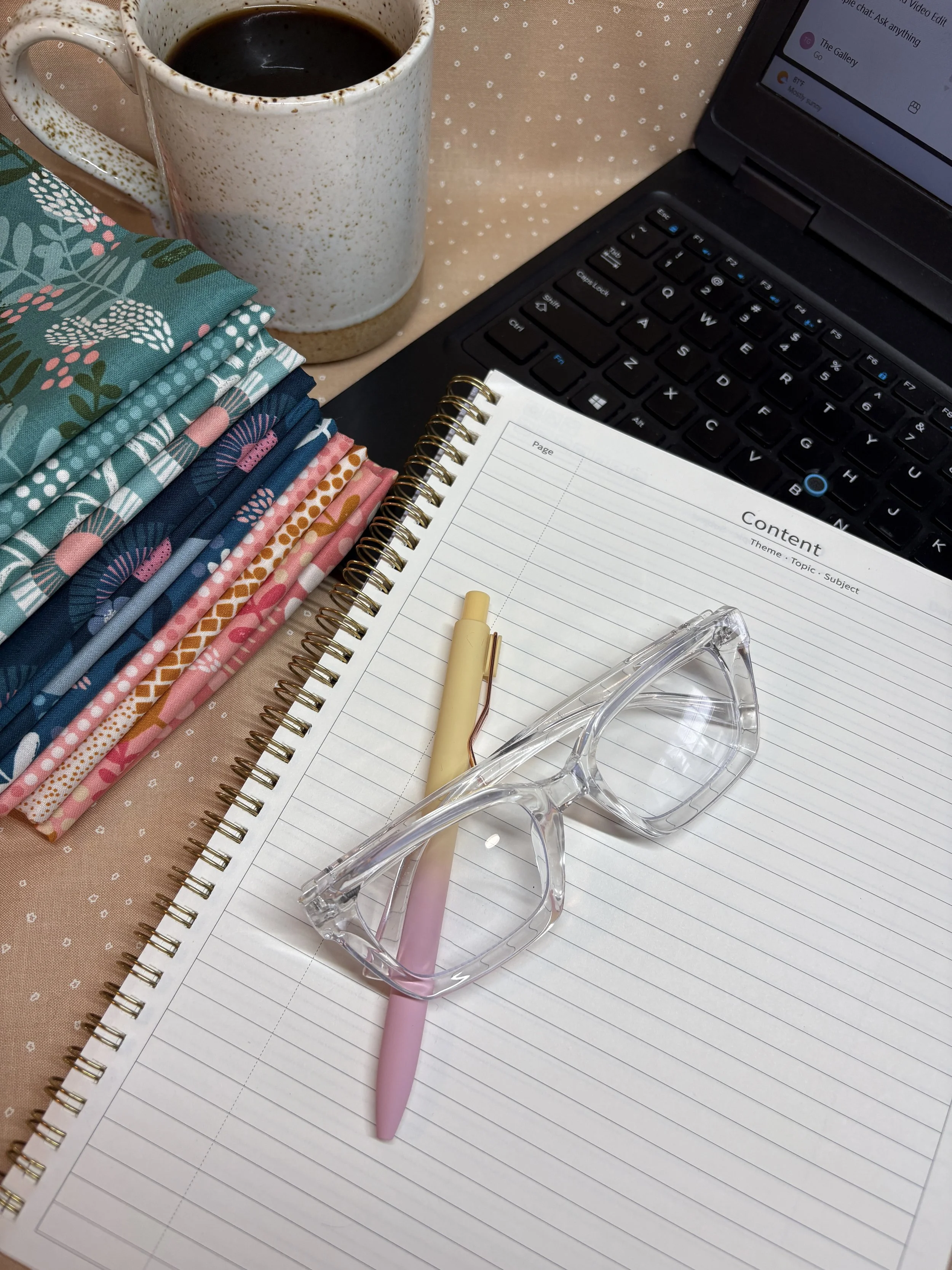 A workspace with a mug of black coffee, a laptop, a spiral notebook, a pink pen, a pair of clear glasses, and stacked colorful fabric pieces on a pink polka-dot fabric surface.