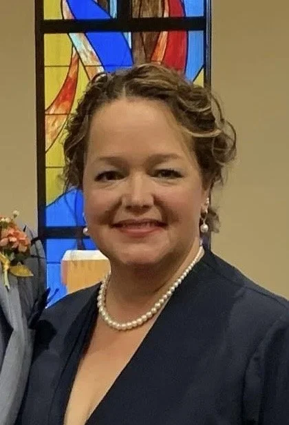 A smiling woman with curly short hair, wearing a dark blazer, pearl necklace, and pearl earrings, standing in front of a colorful stained glass window inside a church.