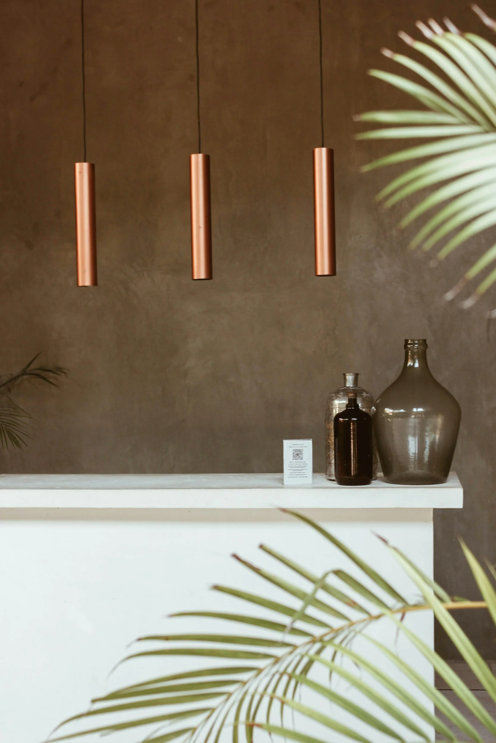 Three copper-colored pendant light fixtures hanging above a white display shelf with glass bottles and a small white stand with text, with a potted plant in the foreground and a brown wall in the background.