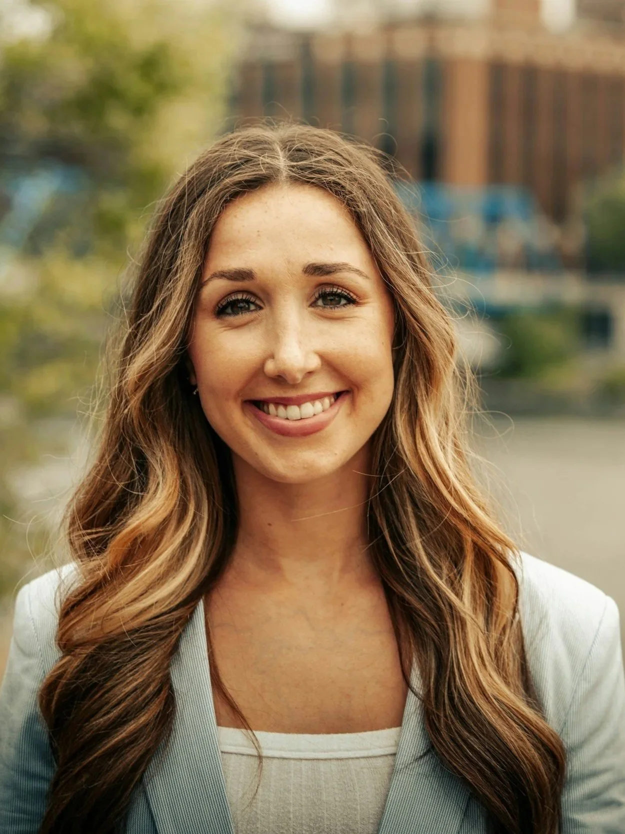 A smiling woman with long wavy brown hair, wearing a light-colored blazer and blouse, standing outdoors with a blurred background of trees and buildings.