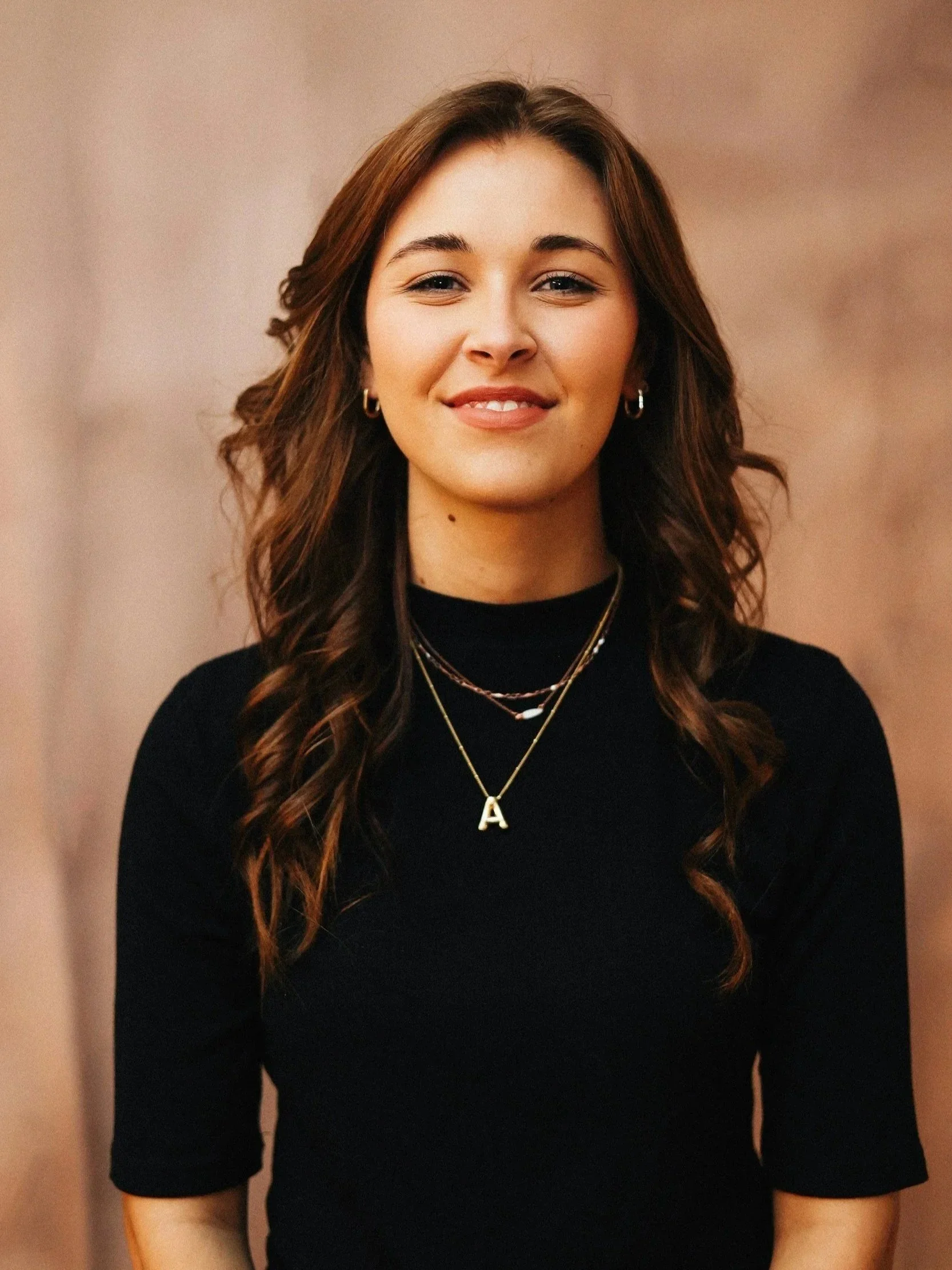 A young woman with long, wavy brown hair, wearing hoop earrings, layered necklaces, and a black long-sleeve top, smiling and standing against a blurred beige background.