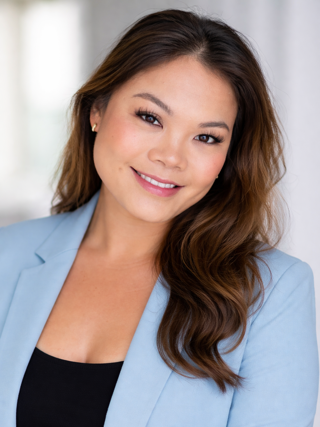 A smiling woman with long, wavy brown hair, wearing a light blue blazer and black top, posing indoors with a blurred background.