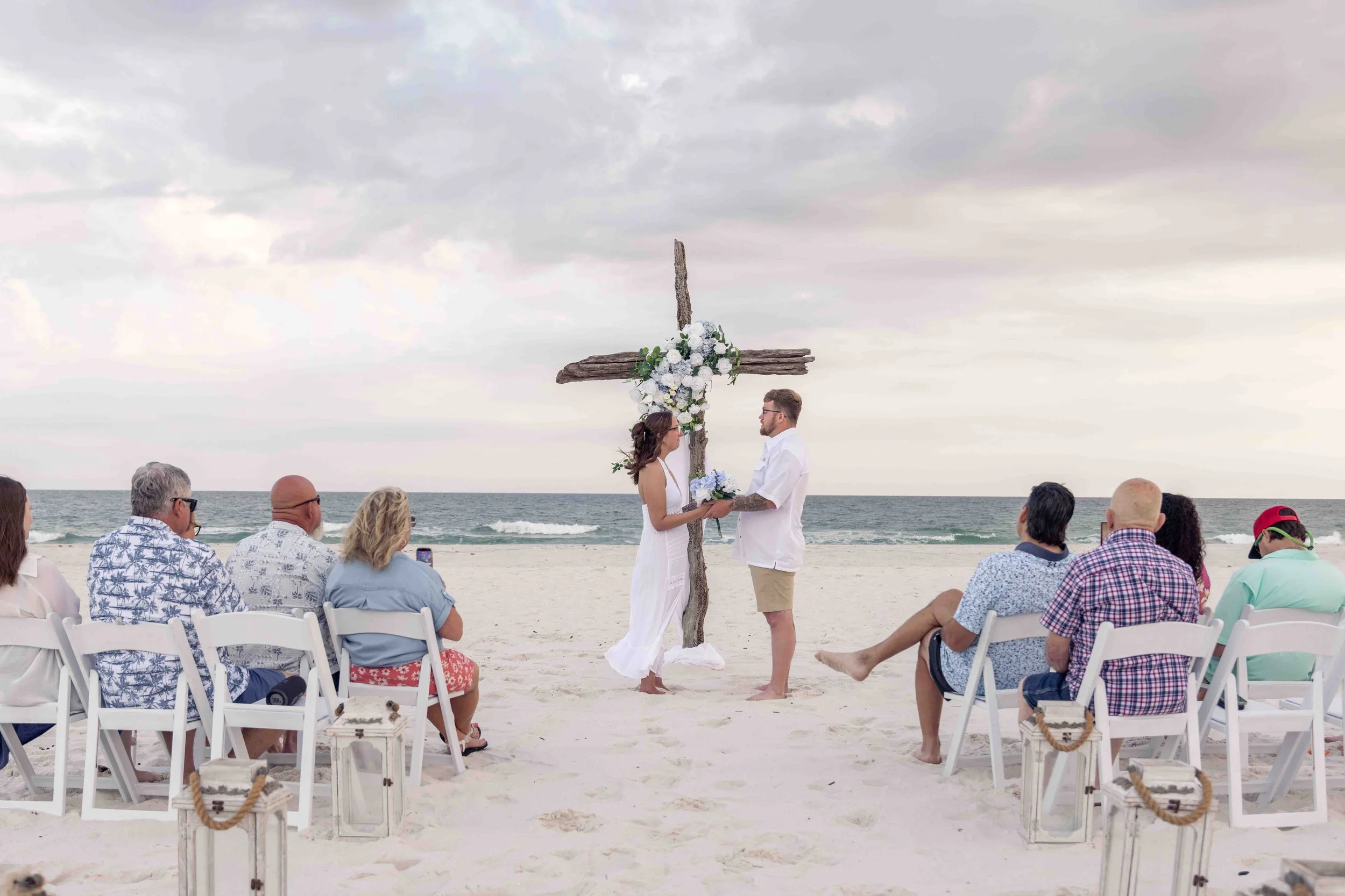 A beach wedding ceremony with a couple standing under a wooden cross decorated with white flowers, surrounded by seated guests on white chairs, with ocean waves and overcast sky in the background.