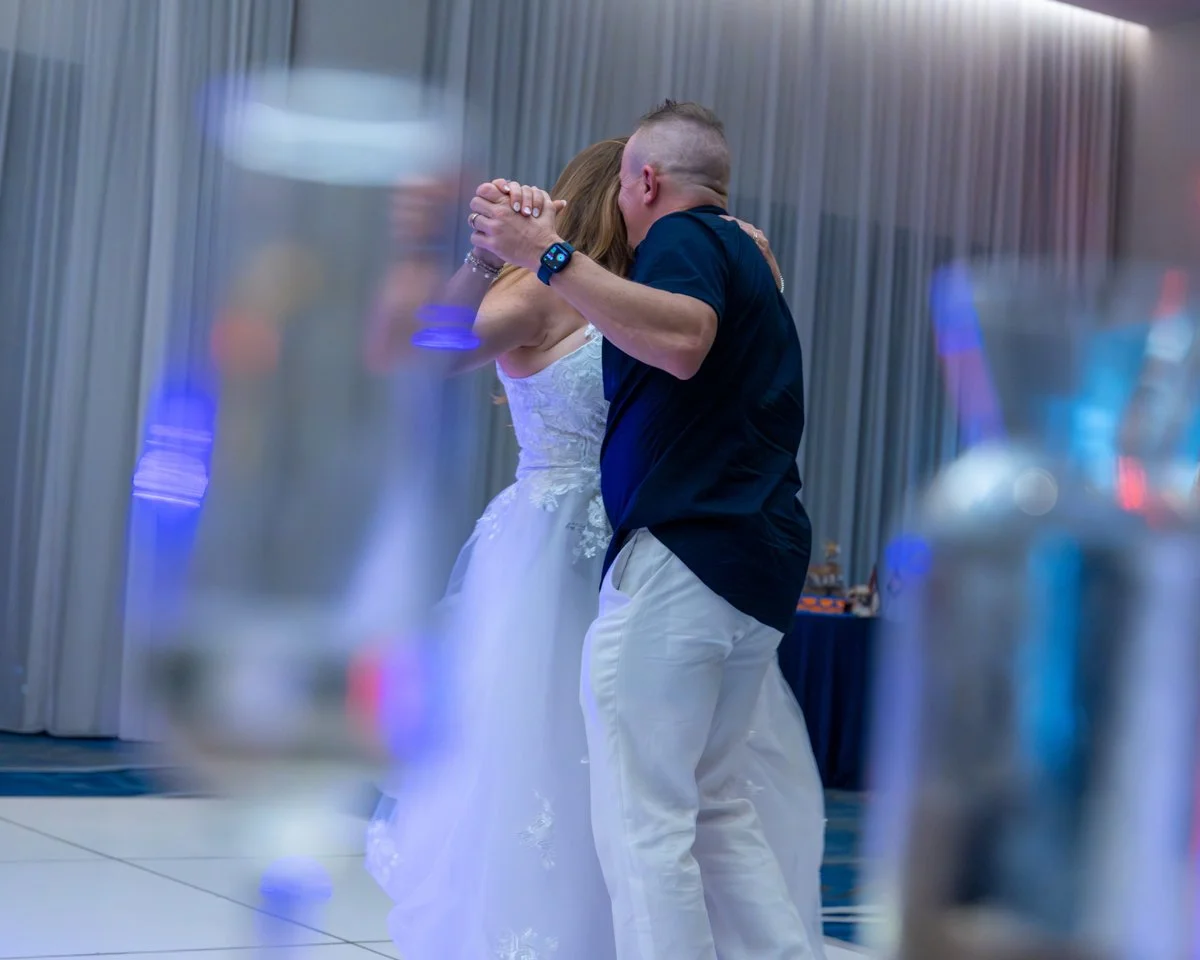 A couple dancing closely at a wedding reception, the woman in a white wedding dress and the man in a dark shirt and light-colored pants, with blurred bottles in the foreground.