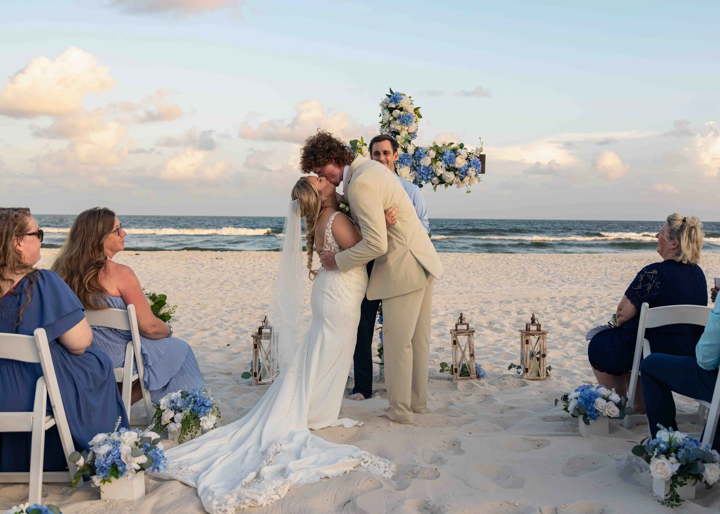 A couple getting married on the beach, surrounded by friends and family, with an ocean and sky in the background. The bride and groom are kissing, with the ocean waves behind them and blue and white floral decorations nearby.