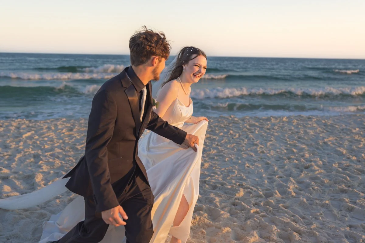 A bride and groom running hand in hand on a beach at sunset, smiling and enjoying their wedding day.
