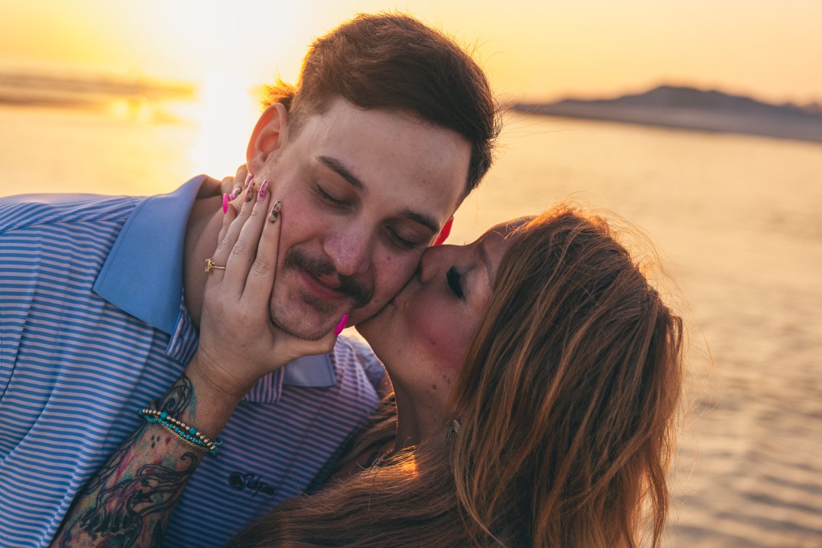 A couple sharing a kiss by the water at sunset, with the man having short dark hair and the woman with long light brown hair.