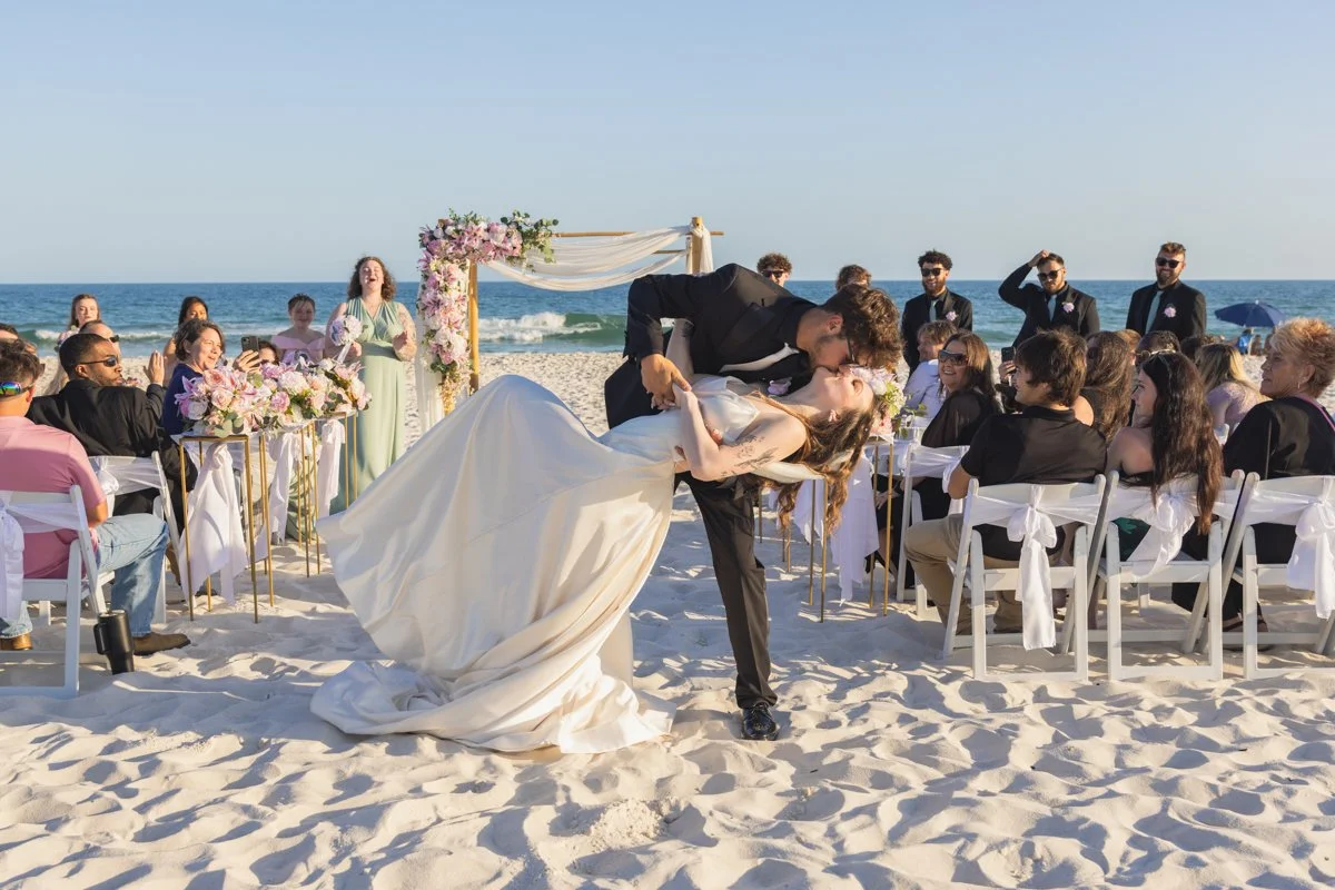 Couple in wedding attire sharing a kiss during their beach wedding ceremony with guests seated on white chairs, a floral arch, and the ocean in the background.