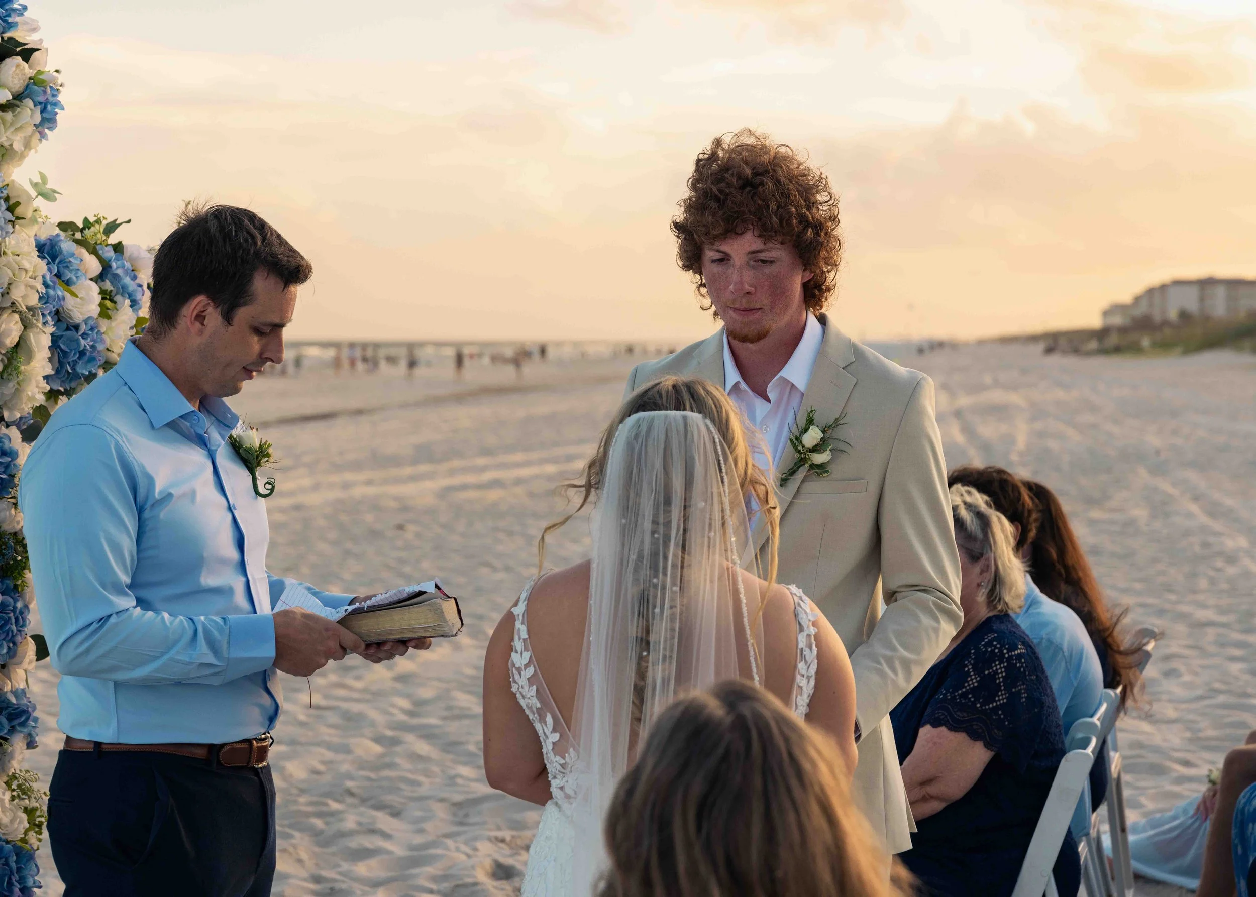 A wedding ceremony taking place on a beach at sunset with the bride, groom, and officiant participating.
