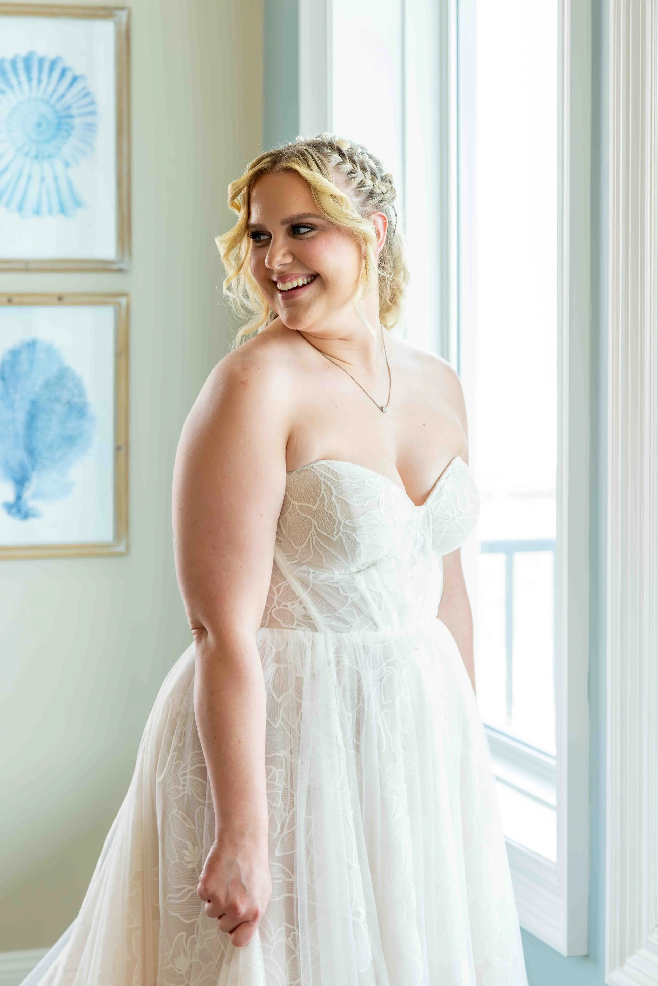 A woman in a white, strapless wedding dress with lace details, standing indoors near windows, smiling and looking to her right.