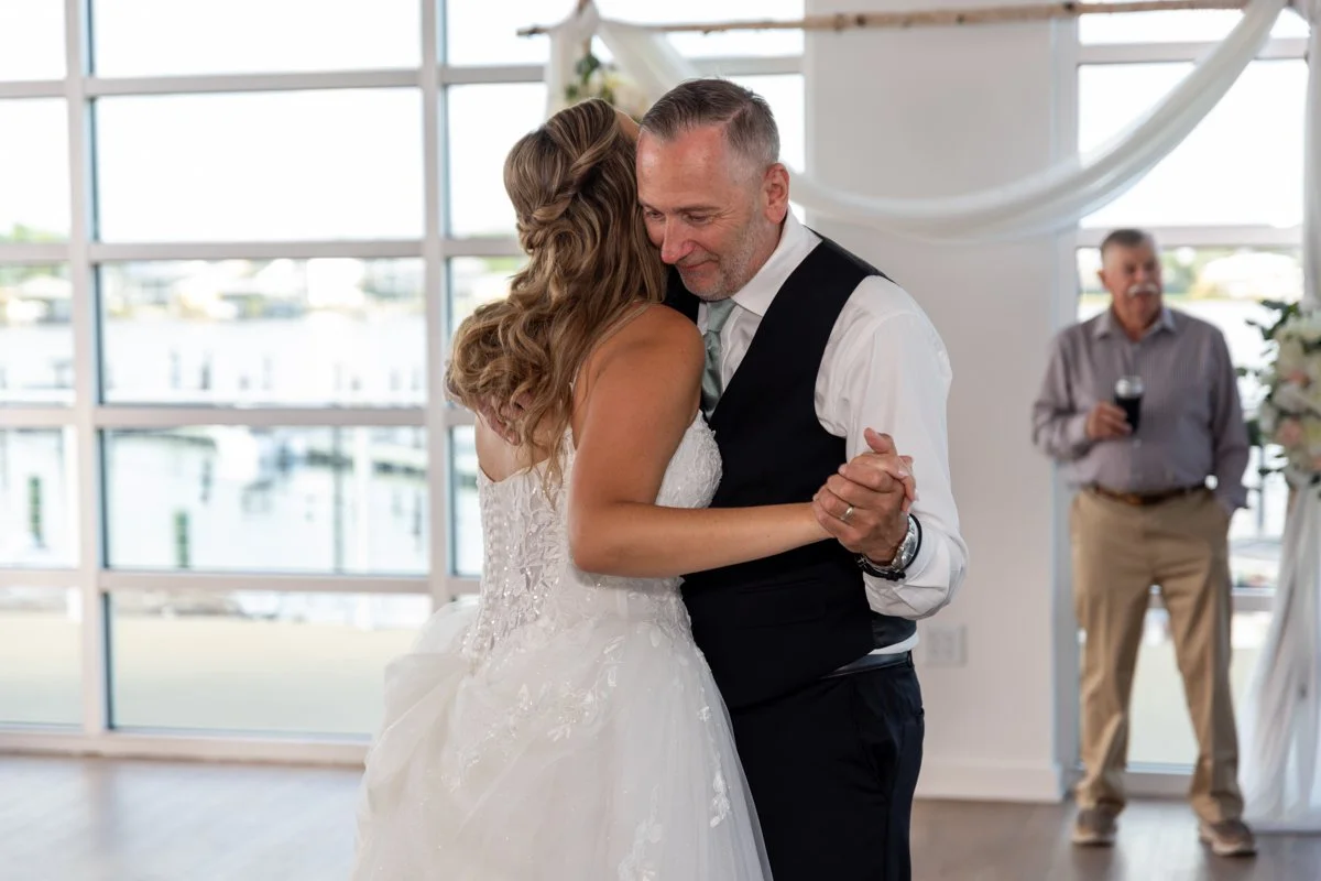 A bride and an older man dancing closely at a wedding, with a man holding a drink in the background near large windows overlooking water.