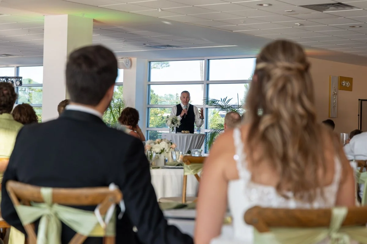 A man and a woman seated at a wedding reception watching a speaker at the podium, with the man in a tuxedo and the woman in a wedding dress, in a brightly lit room with large windows and plants.