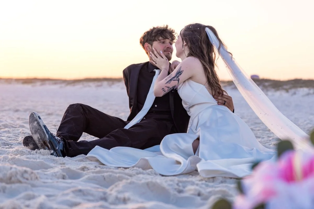 A couple in wedding attire sitting on the sand at the beach during sunset, sharing an intimate moment, with the woman touching the man's face.