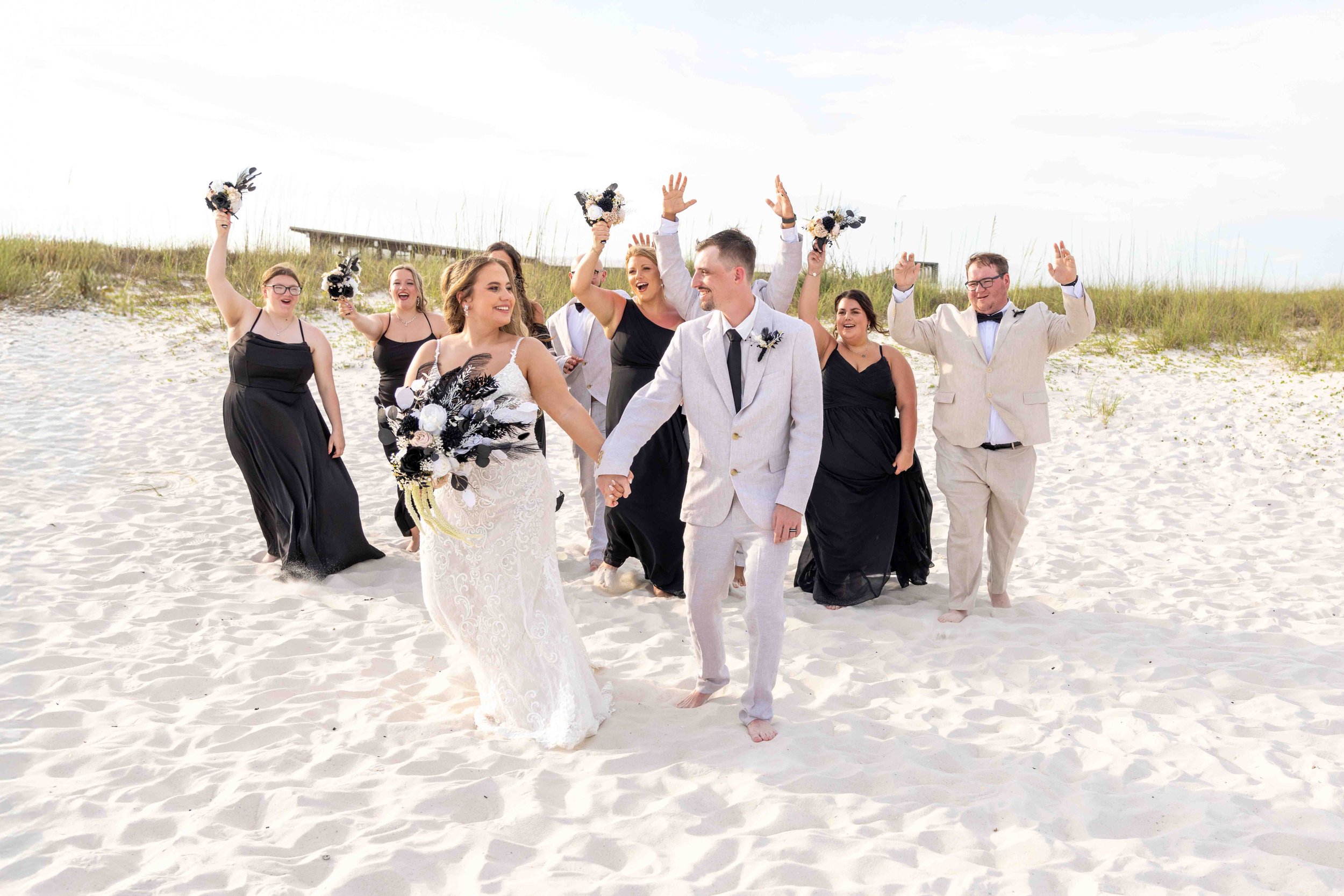 A group of wedding guests and the bride and groom walking barefoot on the beach, with some guests holding bouquets and raising their hands in celebration.
