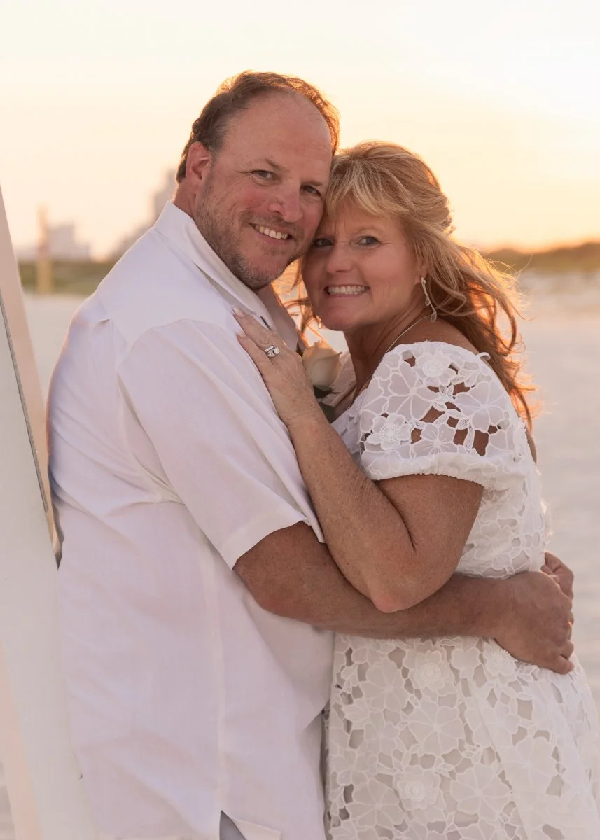 A happy couple embracing on a beach at sunset, with the man wearing a white shirt and the woman in a white lace dress, both smiling warmly.