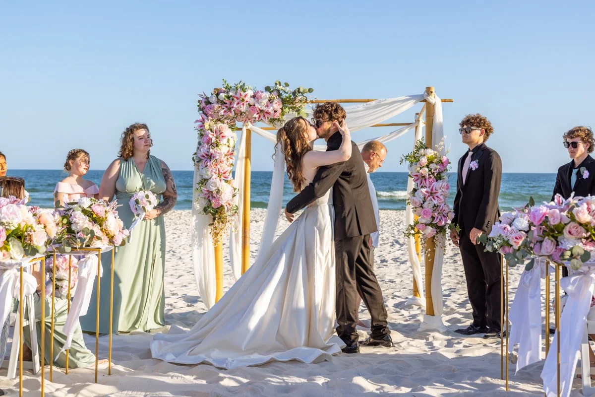 A wedding ceremony on the beach with a couple kissing at the altar, surrounded by friends and family, with ocean and blue sky in the background.