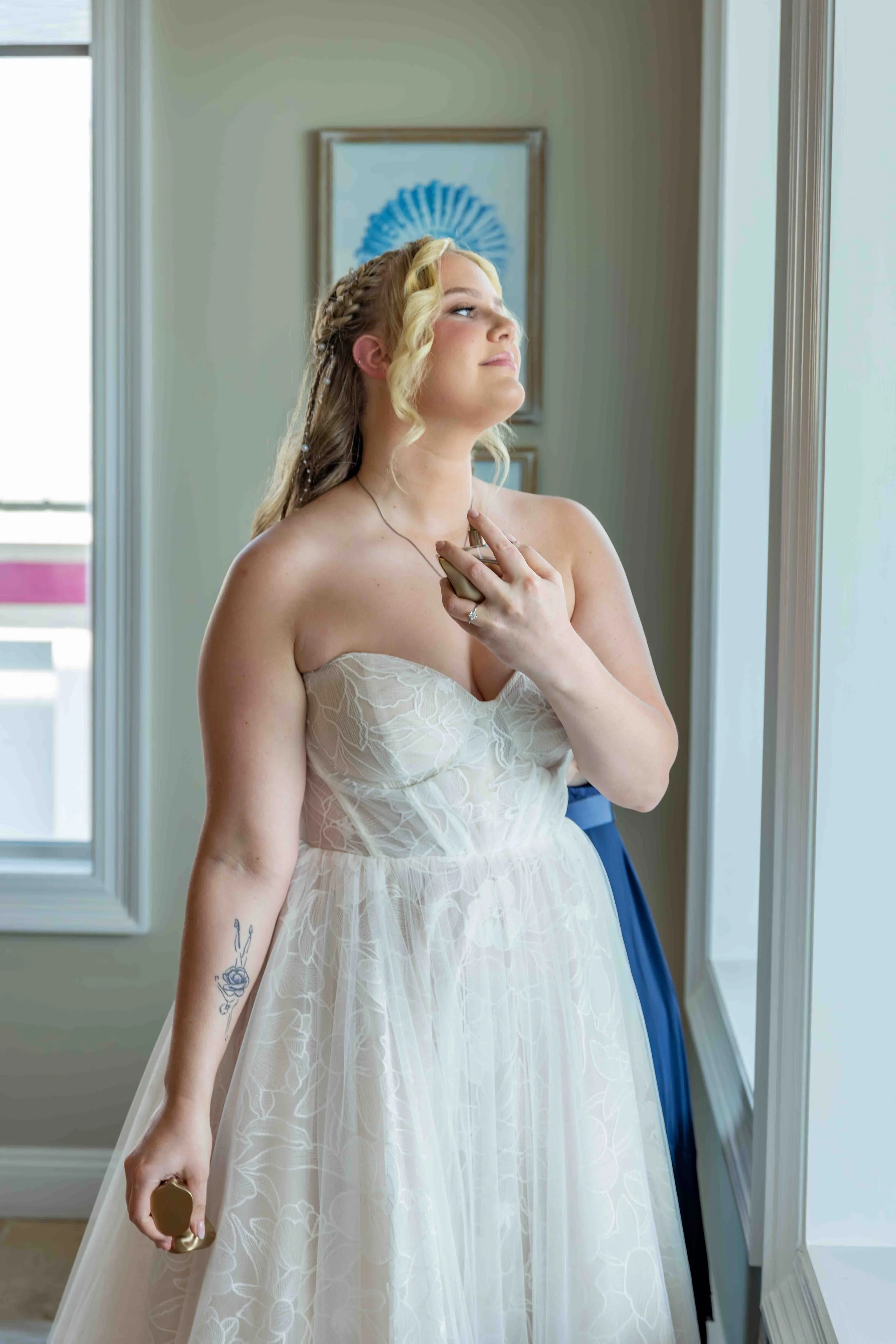 A bride wearing a strapless white wedding dress with floral lace pattern, holding a champagne flute, standing by a window with natural light, with her hand raised near her face, showcasing her tattoo of a rose on her forearm, and a pearl-adorned brai
