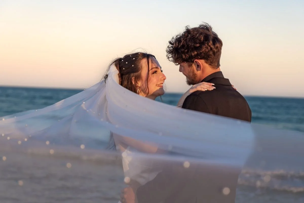 A bride and groom on the beach at sunset, smiling and looking at each other, with the bride's veil flowing in the wind.