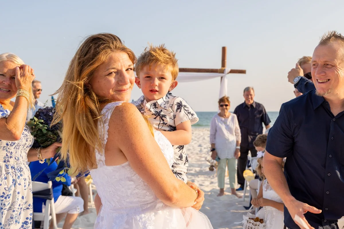 People at a beach wedding, with a woman holding a young boy, a man smiling, and other guests in the background near the ocean and an arch decorated with white fabric.