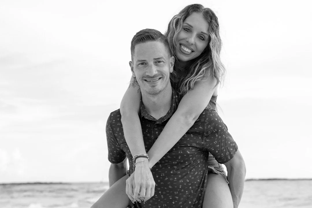A smiling man giving a piggyback ride to a smiling woman at the beach, both looking happy in black and white.