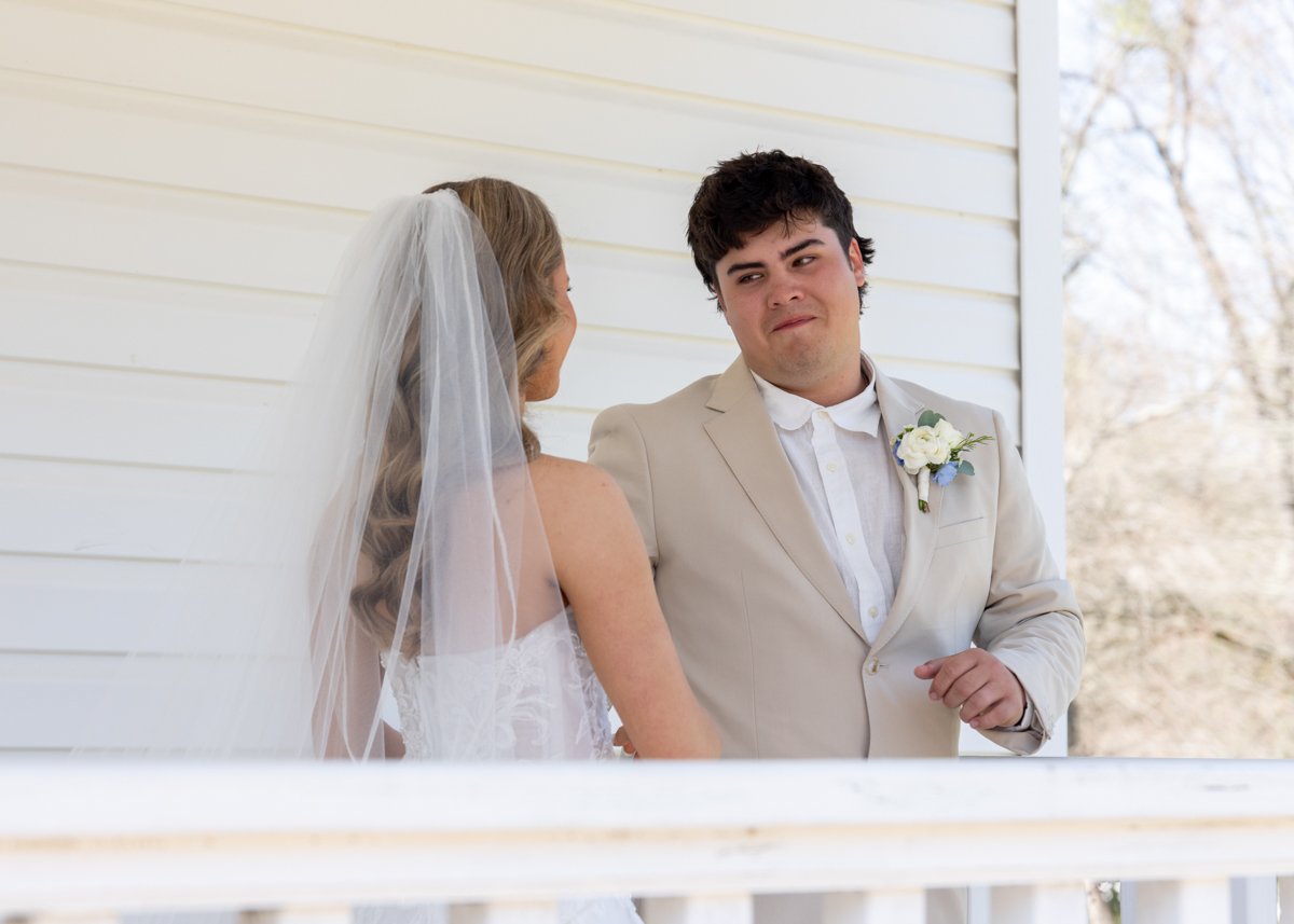 A bride and groom are standing close together on a porch, sharing a moment during their wedding. The bride is wearing a white wedding dress and veil, and the groom is dressed in a light-colored suit with a boutonnière.