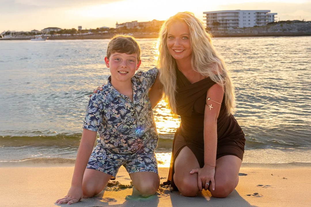 A woman and a boy on a beach at sunset, kneeling on the sand with the ocean and buildings in the background.
