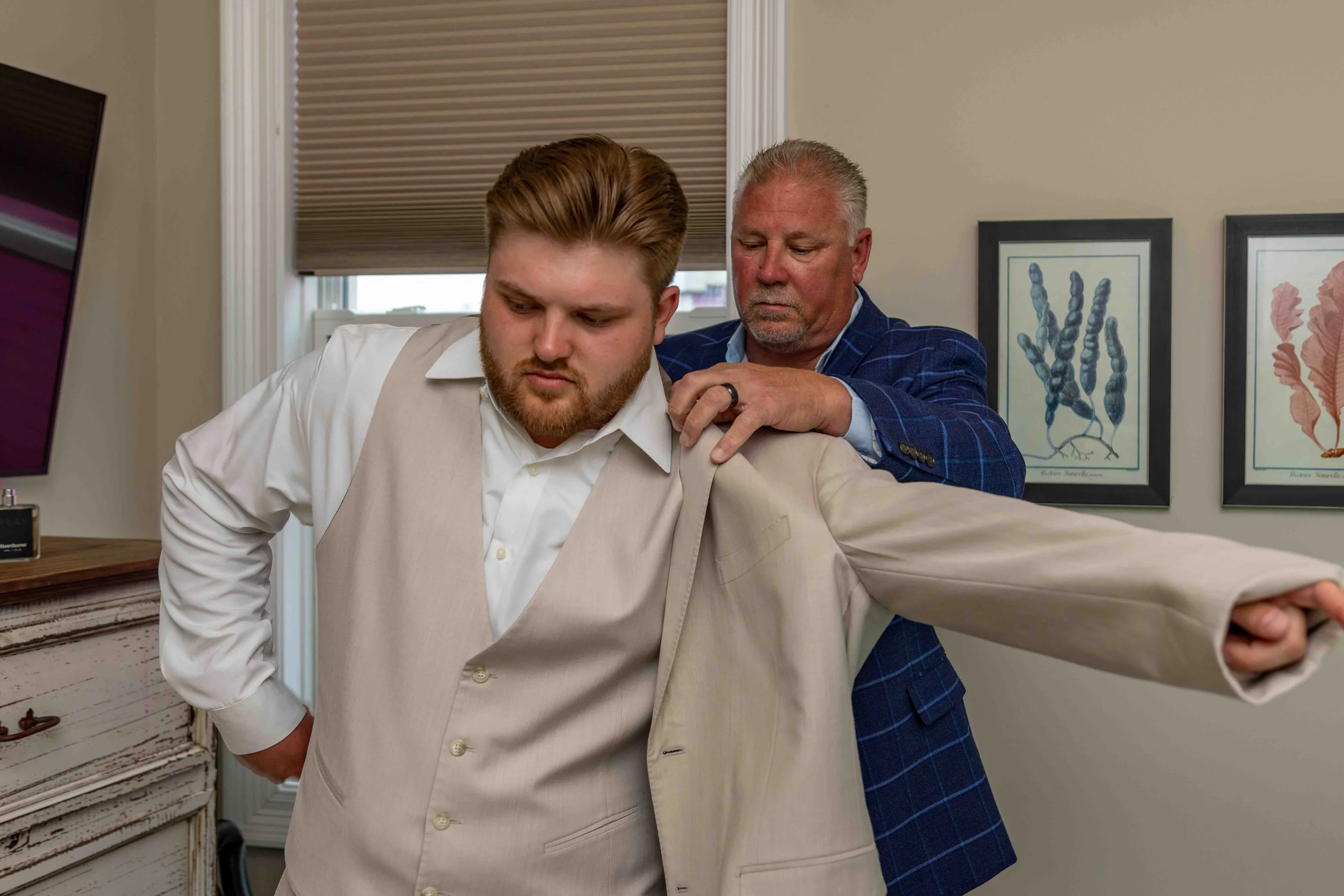 A man with red hair and beard wearing a white shirt and beige vest being adjusted by an older man with gray hair in a blue plaid suit in an office. The older man is fixing the man's shoulder. There are framed pictures on the wall behind them.