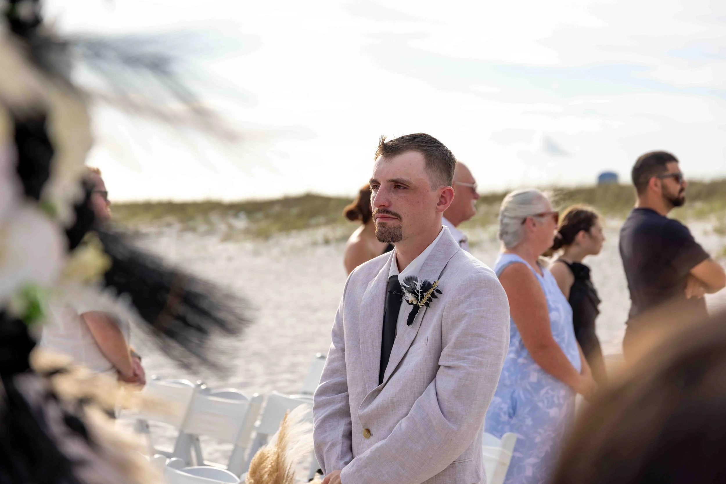 A man with short brown hair, goatee, and mustache wearing a light gray suit with a boutonniere sits with eyes closed at an outdoor wedding ceremony on a beach, surrounded by other guests.