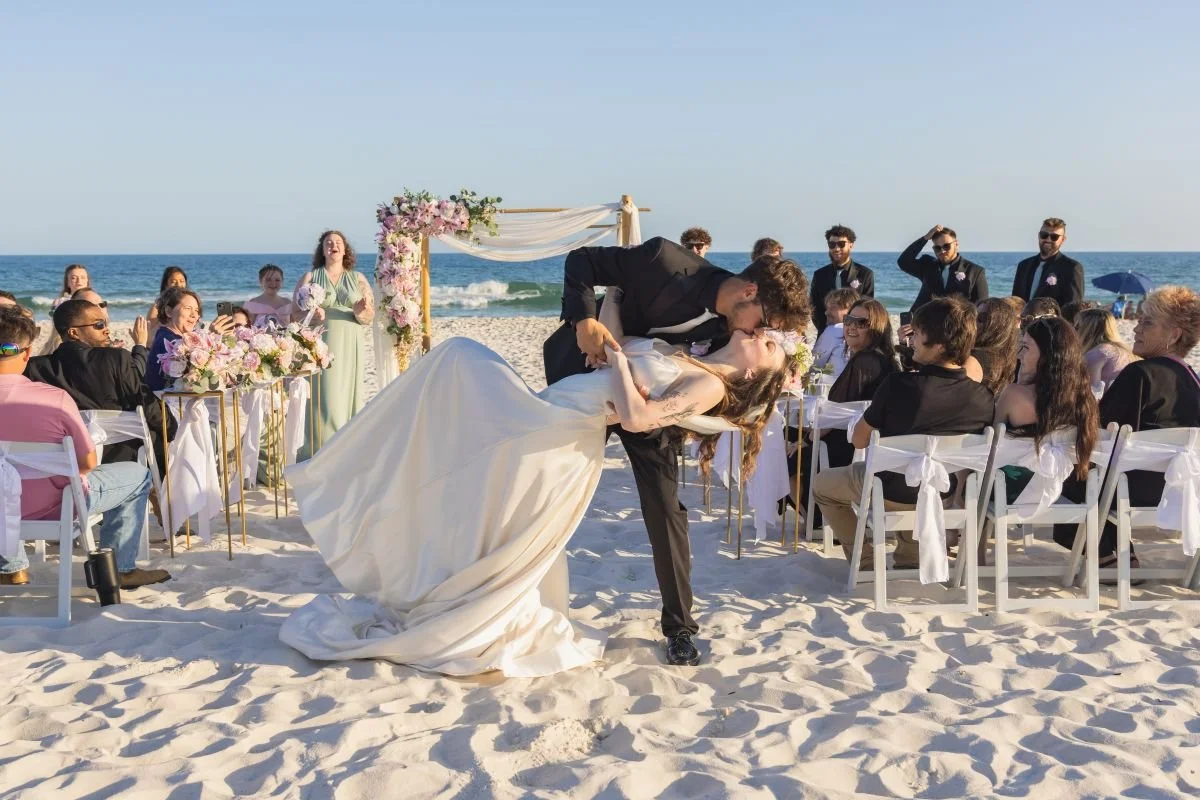A newlywed couple shares a kiss during their beach wedding, with friends and family seated around them and the ocean in the background.