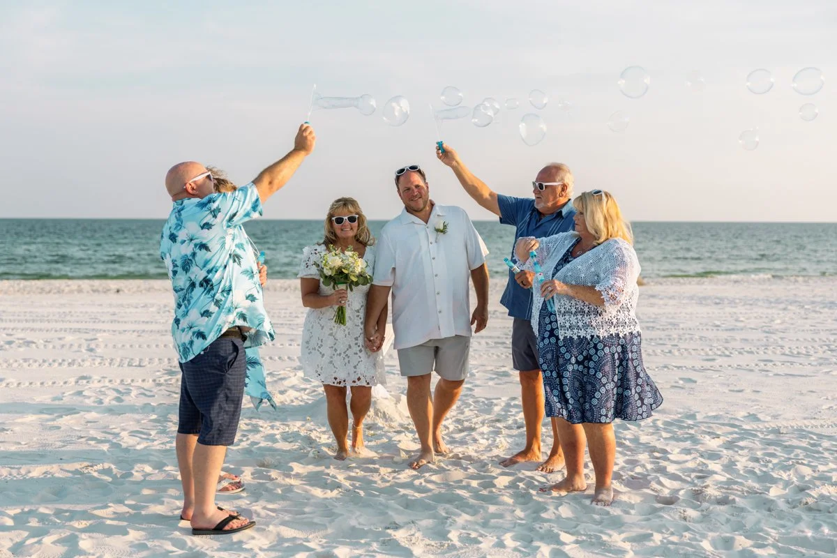 A group of six people at the beach celebrating a wedding, with a bride and groom holding hands in the center, surrounded by friends and family blowing bubbles.