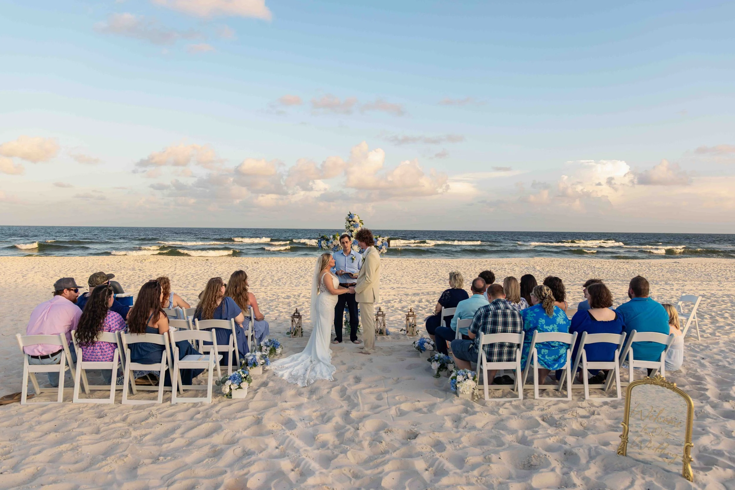 A beach wedding ceremony with a couple exchanging vows, seated guests on white chairs, scenery of the ocean, sky, and clouds, decorated with floral arrangements and lanterns along the aisle.