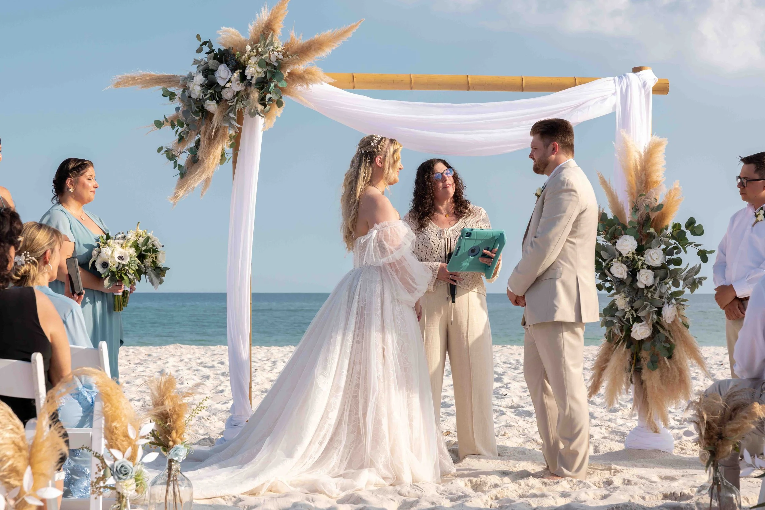 A beach wedding ceremony with a couple standing under a decorated bamboo arch, exchanging vows in front of an officiant, with guests seated nearby. The scene features a bright blue sky, ocean in the background, and floral arrangements with eucalyptus