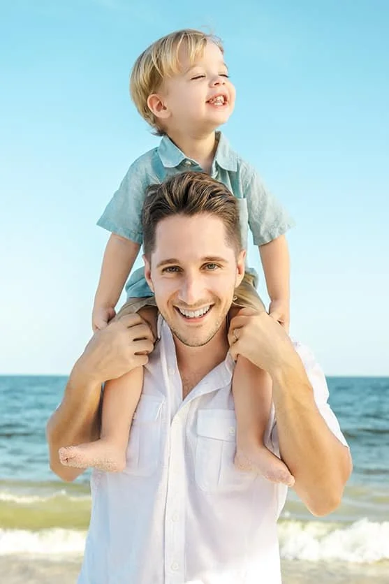 A smiling man carrying a young boy on his shoulders at the beach, with the ocean in the background.