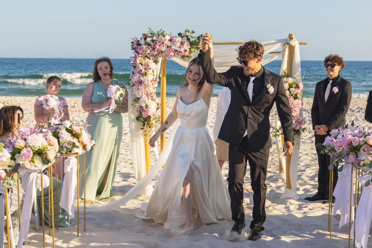 Wedding ceremony on the beach with bride and groom, wedding party, and guests celebrating under a floral arch.