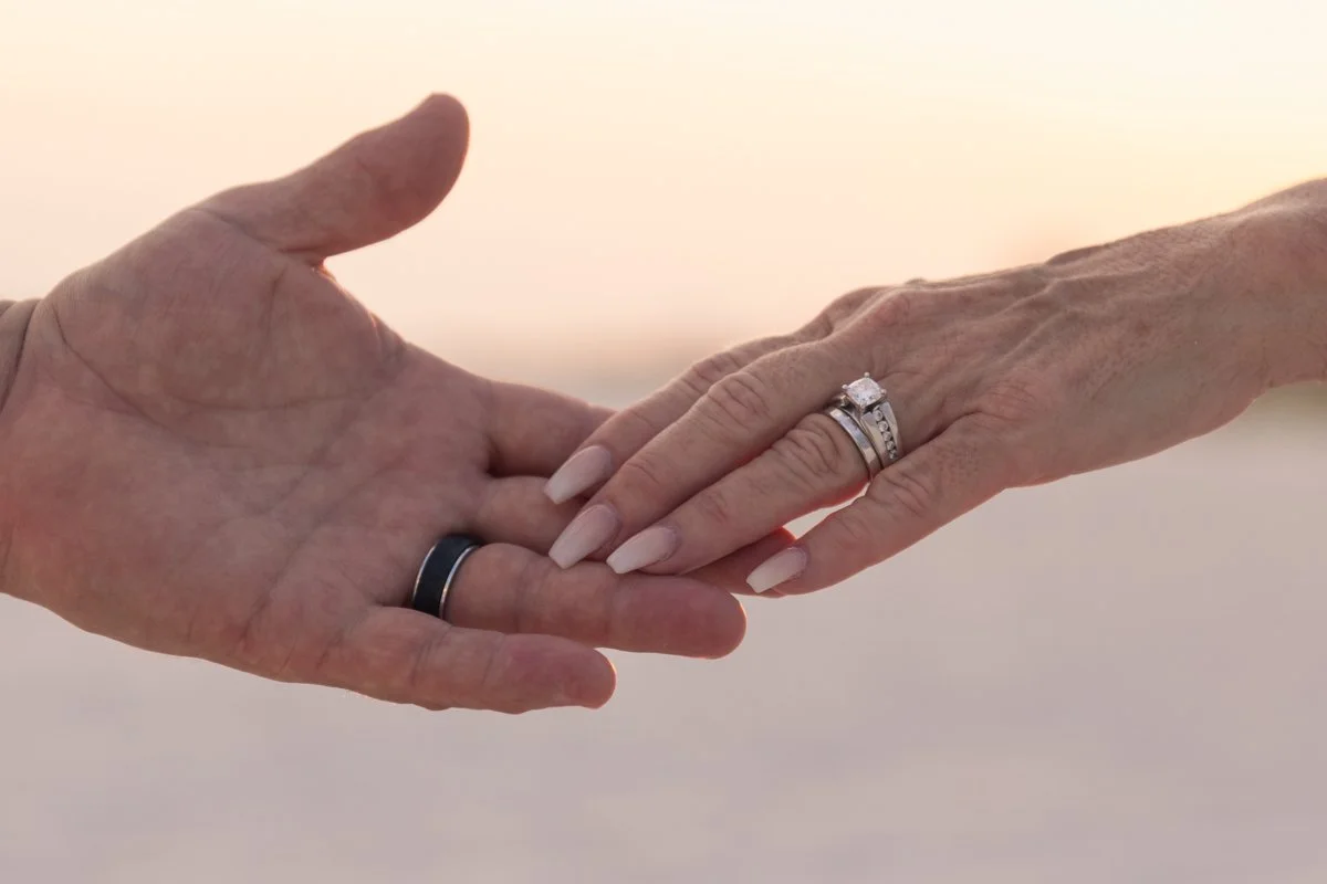 Close-up of two hands reaching out to each other, with wedding rings on their fingers, against a soft background.