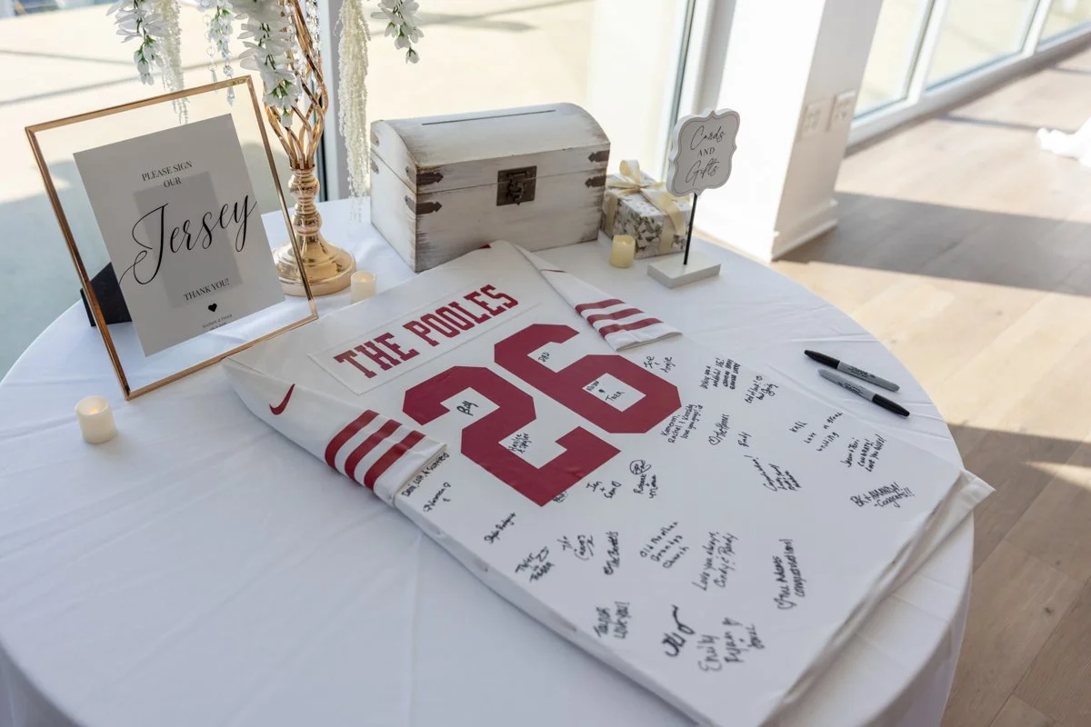 A sports jersey with the number 26 and team name 'The Poles' is displayed on a table, signed by multiple people. The table also has a framed sign, a decorative box, a sign that reads 'Cards and Gifts', a small candle, a gift box, and two black marker