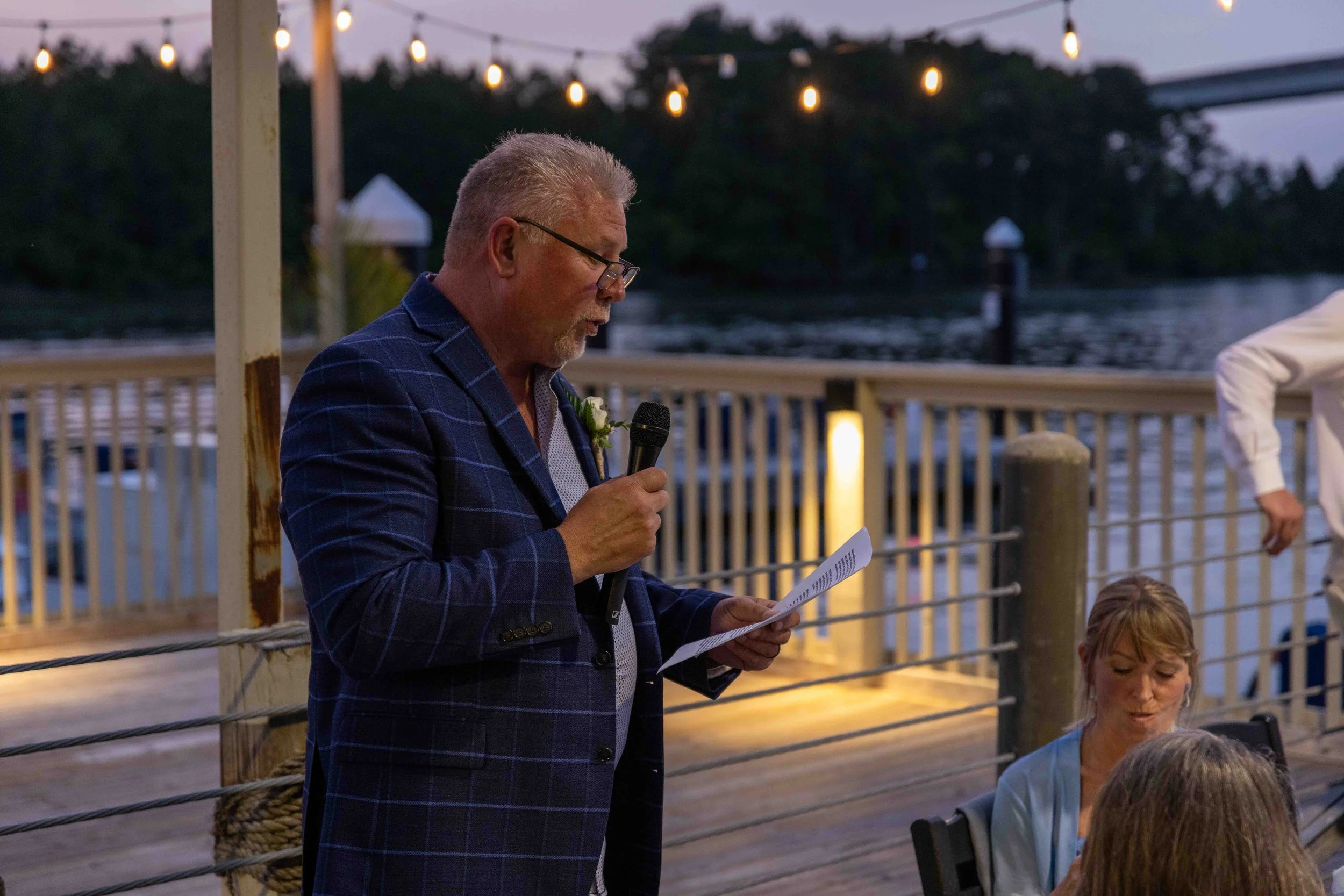 An elderly man with gray hair and beard, wearing glasses and a blue checkered suit, is holding a microphone and reading from a paper at an outdoor evening event near a body of water, with string lights overhead and a wooden railing.