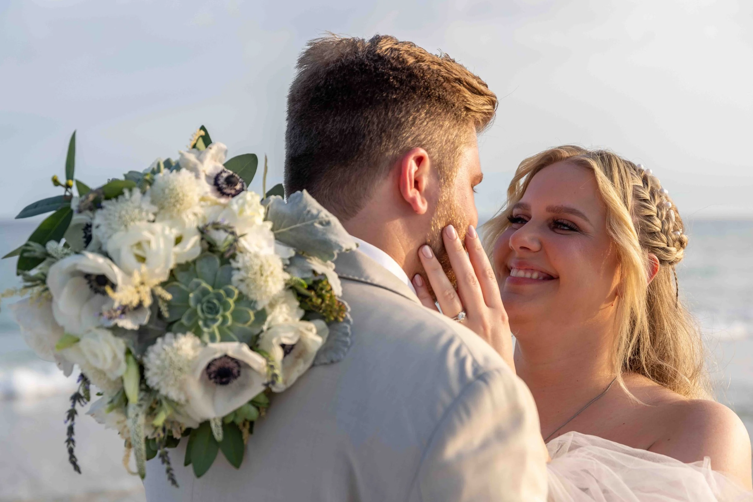 A bride and groom at their wedding, embracing each other on the beach during sunset, with the groom holding a bouquet of white flowers.