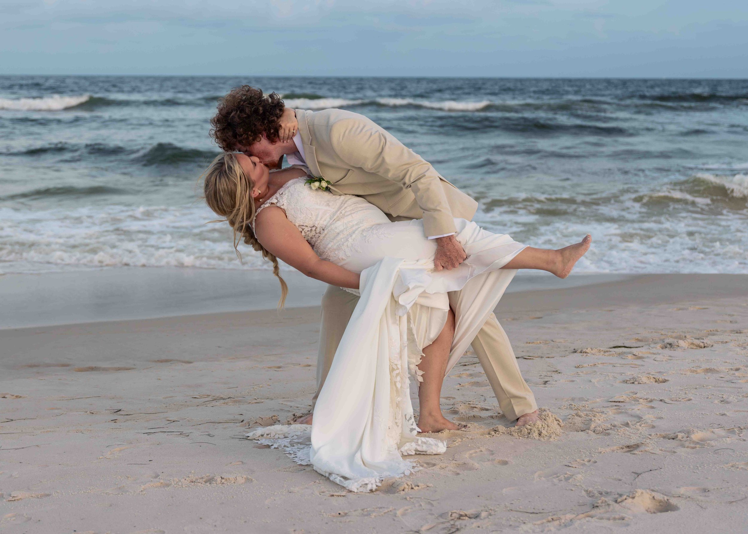 A bride and groom on a beach, with the groom dipping the bride in a romantic kiss, both dressed in wedding attire.