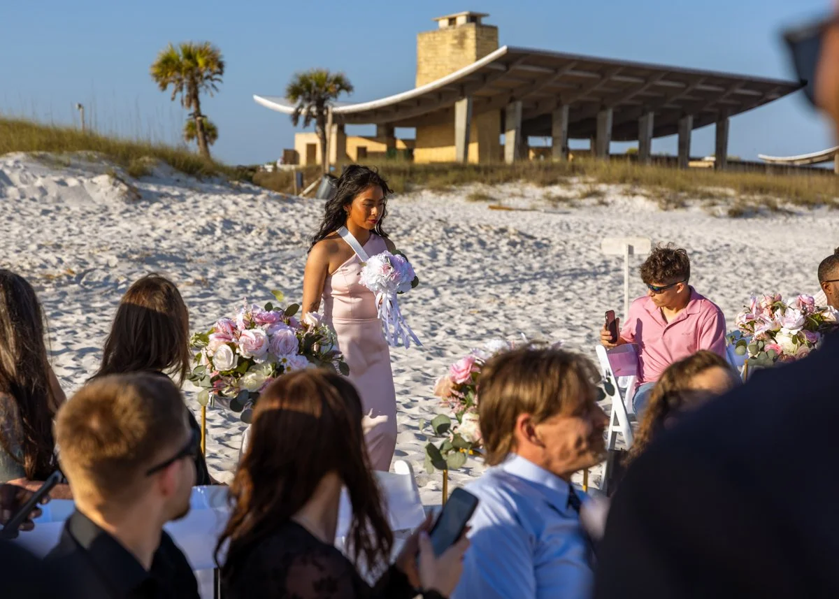A woman in a pink dress holding a bouquet walking on a beach during a wedding ceremony, with seated guests and flowers, and a building with palm trees in the background.