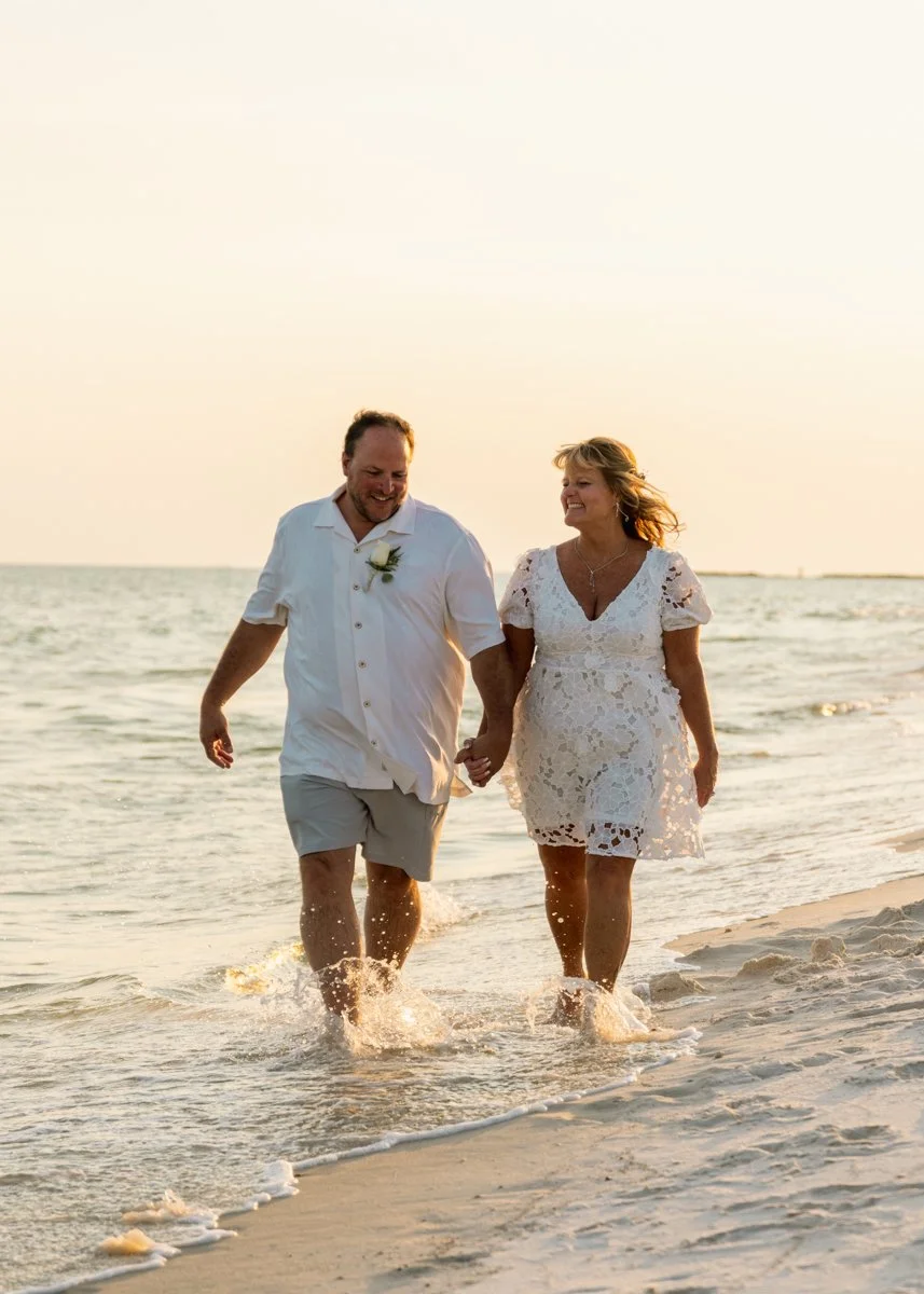 A couple is walking hand-in-hand along the shoreline at the beach during sunset, dressed in white with the woman wearing a lace dress and the man in a white shirt and shorts, smiling and enjoying the moment.