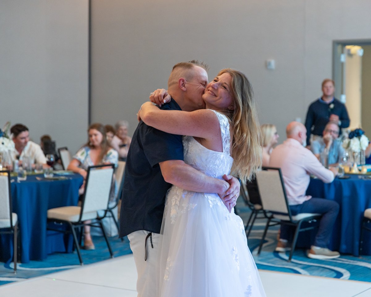 A couple dancing and hugging at a wedding reception, with guests seated at tables in the background.