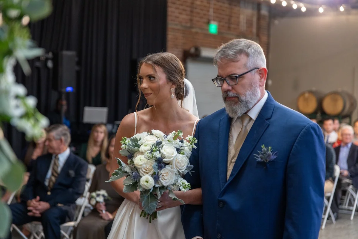 A bride holding a bouquet of white and purple flowers walking down the aisle with her father during a wedding ceremony in a rustic indoor venue.