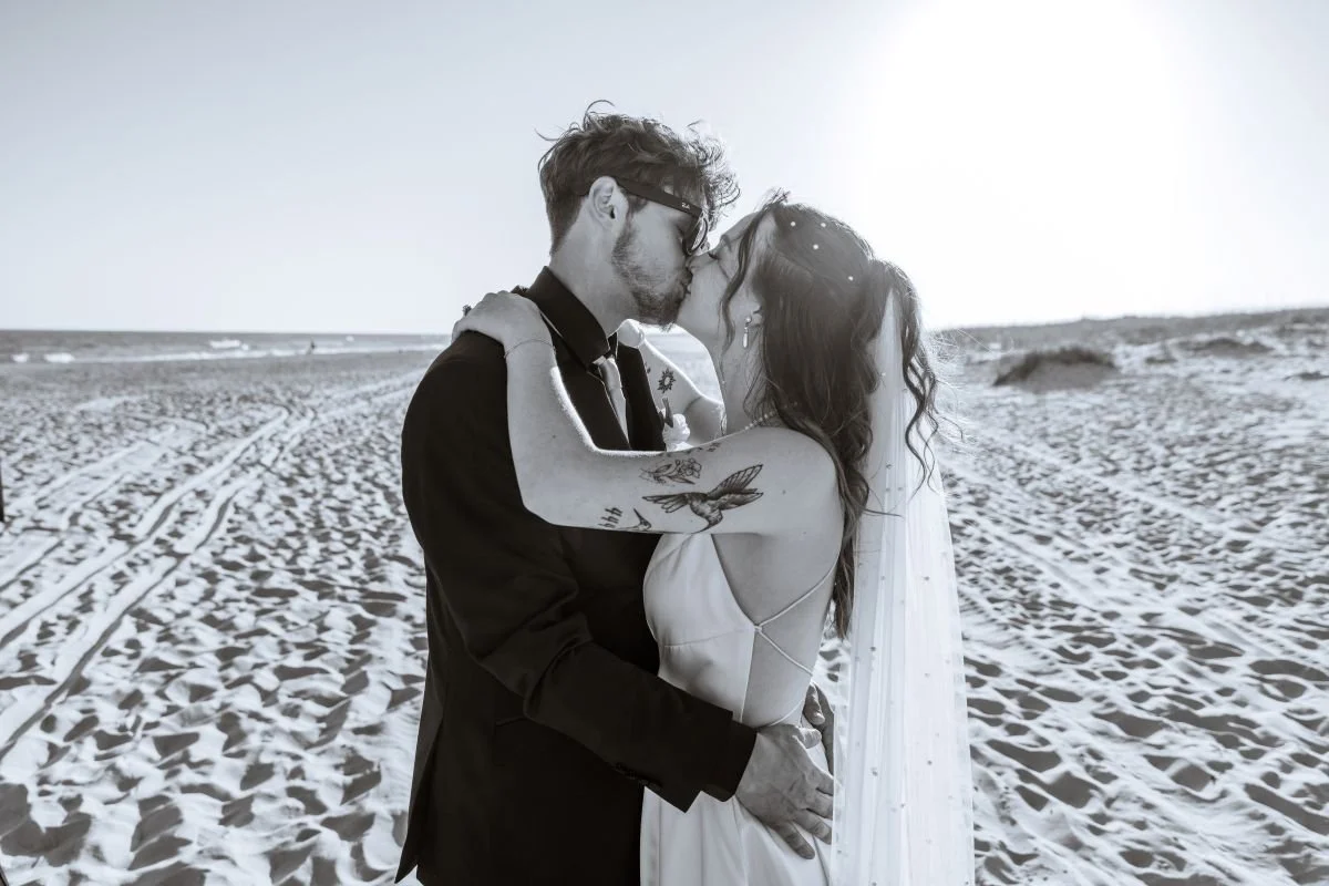 A black and white photo of a couple kissing on a beach, with sandy terrain and the ocean in the background.